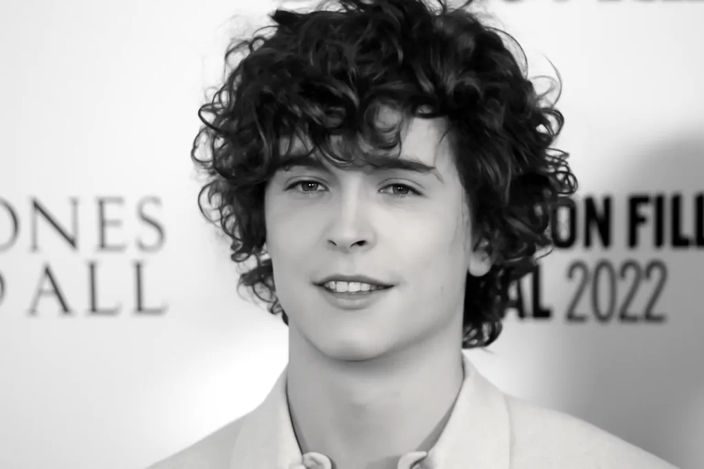 "A black-and-white image of Timothy Shalloway smiling on the red carpet, his curly hair framing his face against a blurred event backdrop